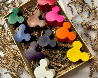 May include: A box of colorful, Mickey Mouse-shaped crayons in various colors, including red, pink, orange, yellow, green, blue, and white. The crayons are arranged in a rectangular box filled with brown crinkle paper.