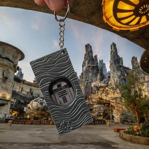 May include: A silver rectangular keyring featuring R2-D2. The keyring has a textured wave pattern and is held up in front of a Star Wars-themed backdrop, including a building and rock formations.