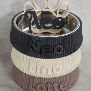 May include: A personalized pet bowl with a stainless steel interior and a stacked, ribbed exterior in black, cream, and brown. The name "Neo" is displayed in black, with a paw print accent. The names "Lino" and "Lotte" are also visible.