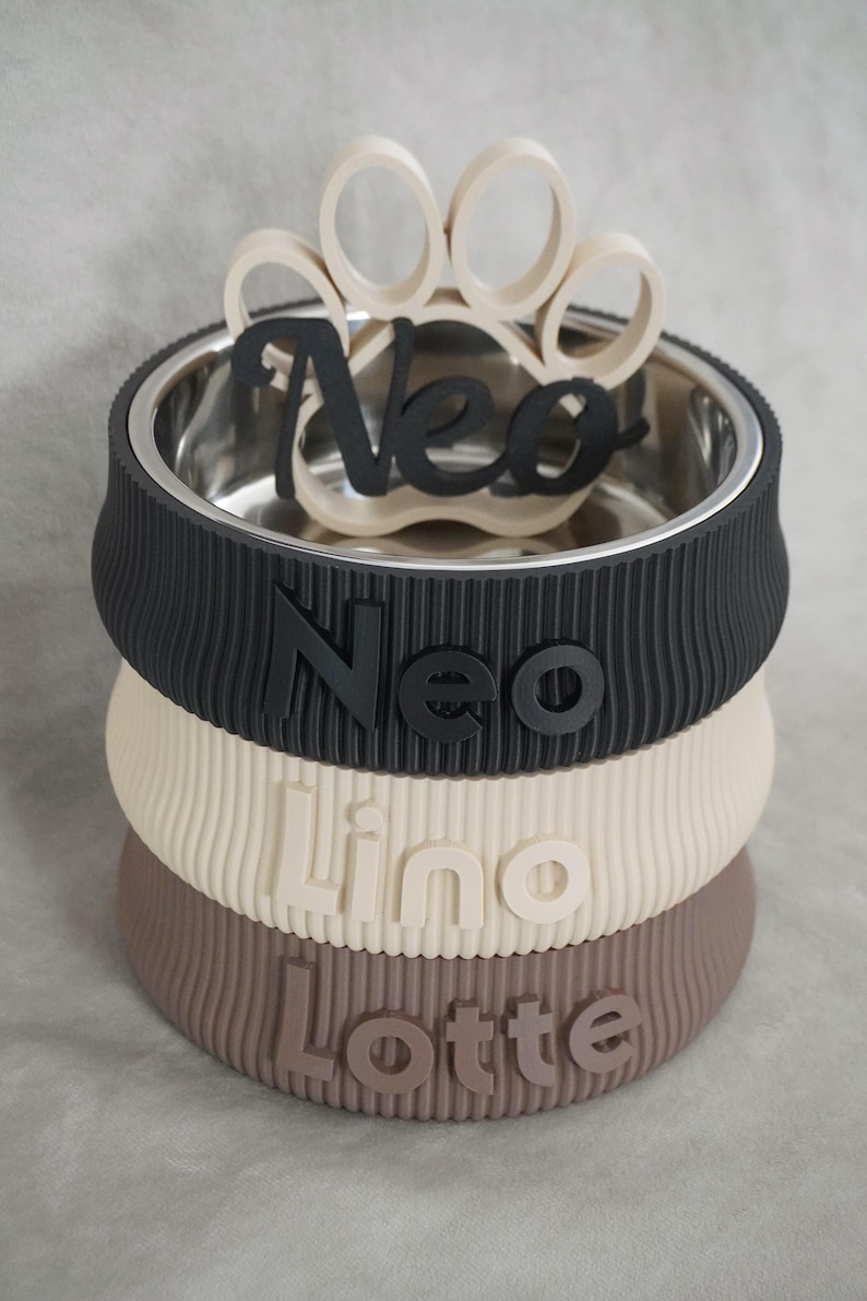 May include: A personalized pet bowl with a stainless steel interior and a stacked, ribbed exterior in black, cream, and brown. The name "Neo" is displayed in black, with a paw print accent. The names "Lino" and "Lotte" are also visible.