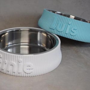May include: Two pet bowls with stainless steel interiors. One is white with the name "Ernie" embossed, the other is light blue with "Luis" embossed. Both bowls have a ribbed exterior design.