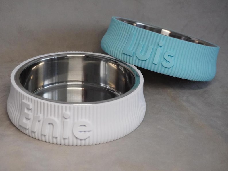 May include: Two pet bowls with stainless steel interiors. One is white with the name "Ernie" embossed, the other is light blue with "Luis" embossed. Both bowls have a ribbed exterior design.