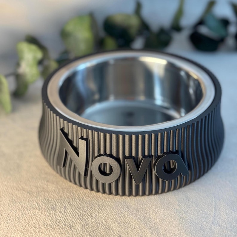 May include: A black pet bowl with a stainless steel interior. The bowl has a ridged exterior with the name "Nova" in raised letters. The bowl is designed for pets and is likely used for food or water.