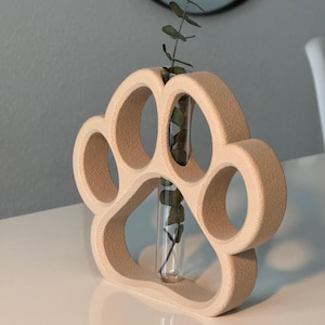 Peut inclure: Un vase beige en forme de patte avec un tube en verre transparent au centre, contenant des brins verts. Le vase est fait d'un matériau texturé et repose sur une surface blanche. Un objet décoratif unique.