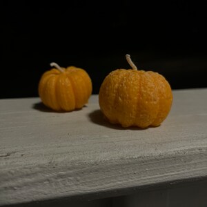 Puede incluir: Dos peque&ntilde;as velas de color naranja en forma de calabaza con mechas blancas sobre una superficie de madera blanca.