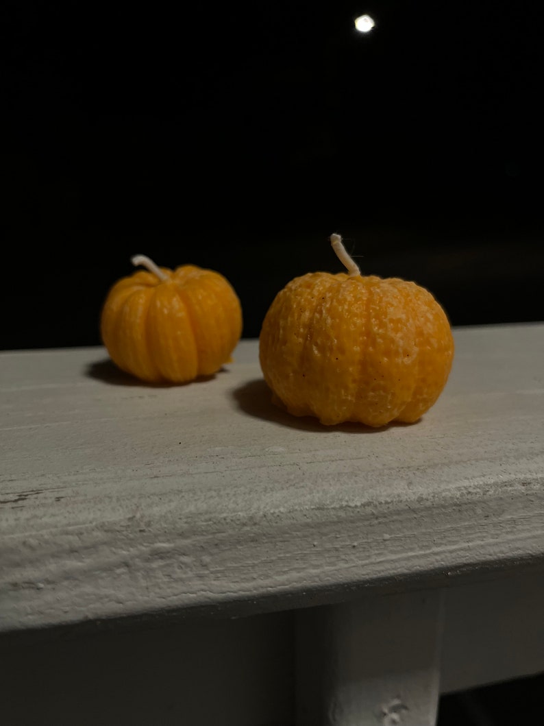 Puede incluir: Dos peque&ntilde;as velas de color naranja en forma de calabaza con mechas blancas sobre una superficie de madera blanca.