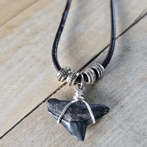 May include: A shark tooth pendant necklace. The pendant is a dark gray shark tooth wrapped in silver wire, suspended from a black leather cord. Silver beads accent the cord near the clasp. The necklace rests on a wooden surface.