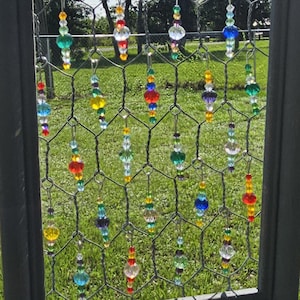May include: A decorative hanging art piece featuring a dark gray frame with chicken wire. Colorful glass beads in various shapes and sizes, including red, blue, green, and yellow, are suspended from the wire, creating a vibrant display.