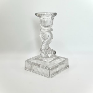May include: A clear glass candlestick with a square base and a decorative fish-shaped stem.