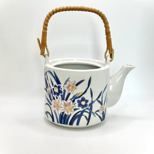 Può includere: Teiera in ceramica bianca con manico in bambù e design floreale. La teiera presenta un motivo dipinto di fiori rosa, blu e gialli con foglie verdi. Il manico è in bambù intrecciato.