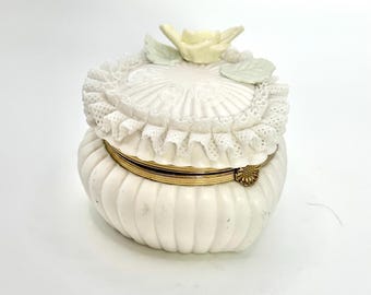 May include: White ceramic trinket box with a hinged lid. The lid is decorated with a white lace trim and a yellow flower with green leaves. The box has a ribbed design.