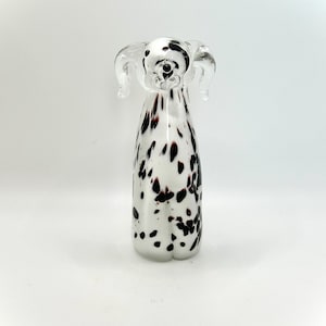 Op de afbeelding: Een helder glazen Dalmatische hondenfiguur met zwarte en bruine vlekken. De hond heeft grote, slappe oren en een gedetailleerd gezicht. Het beeldje staat rechtop op een wit oppervlak.