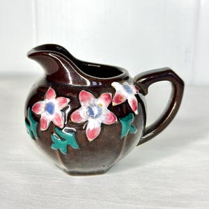 Może przedstawiać: Mały brązowy dzbanek do śmietanki z ceramicznym wzorem kwiatowym. Dzbanek ma zaokrąglony korpus z uchwytem i dzióbkiem. Kwiaty są różowe, białe i niebieskie z zielonymi liśćmi.