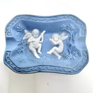 May include: A blue ceramic ashtray featuring a white raised design of two cherubs, one playing a musical instrument. The ashtray has a decorative border.