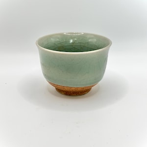 May include: A celadon-glazed ceramic teacup with a crackle finish. The cup has a light green hue with a brown base. The cup is a simple, elegant design.