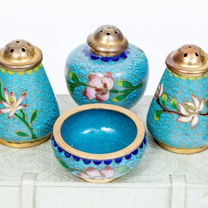 May include: A collection of four decorative cloisonné items. The set comprises two salt shakers, a pepper shaker, and a small bowl. The pieces are turquoise with floral patterns and gold-coloured metal accents. The set is presented on a white surface.