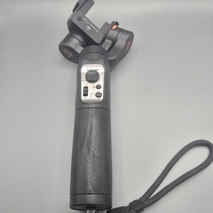 Pode incluir: Um estabilizador de gimbal portátil preto com punho texturizado, alça de pulso e painel de controle com joystick, botões e interruptores. O dispositivo apresenta a marca Hohem e o modelo Steady Pro4 na carcaça do motor.