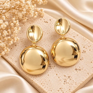 May include: A pair of gold-toned drop earrings featuring a polished, smooth finish. Each earring consists of a smaller circular top disc connected to a larger, rounded circular pendant below, creating a reflective, metallic, and layered design.