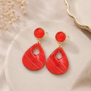 May include: A pair of drop earrings featuring a circular red stud top connected to a larger, teardrop-shaped red pendant with a cutout center. The red material has a subtle, swirling marbled texture, and the hardware is a polished gold tone.