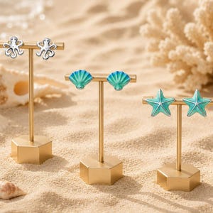 May include: Three pairs of stud earrings displayed on gold-toned T-bar stands. One pair features silver-toned octopuses, one pair features blue and green gradient scallop shells, and one pair features blue and green gradient five-pointed starfish.