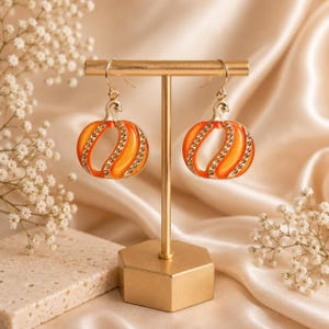 May include: A pair of gold-toned dangle earrings shaped like pumpkins. Each earring features a bright orange enamel center with vertical gold-toned ribs accented by small, clear, sparkling crystals along the curved edges of the pumpkin design.