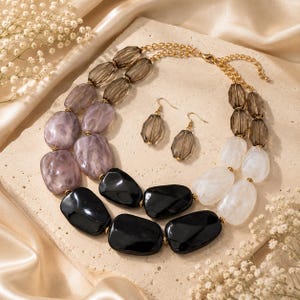 May include: A multi-strand statement necklace and matching drop earrings set. The necklace features large, smooth, irregular beads in shades of black, white, and marbled lavender, accented by translucent smoky gray faceted beads and gold-tone metal chains.