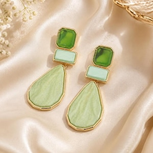 May include: A pair of green and gold earrings. Each earring features a green square at the top, a rectangular green stone in the middle, and a large, teardrop-shaped green stone at the bottom. The stones are set in gold-colored metal.
