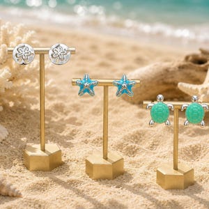 May include: Three gold-toned T-bar earring stands displaying pairs of earrings. The left stand holds silver sand dollar earrings, the center stand holds blue starfish earrings, and the right stand holds green sea turtle earrings.