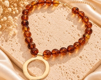May include: A necklace featuring a strand of translucent, round, amber-colored beads and a large, polished gold-tone circular pendant. The necklace includes a gold-tone metal chain extension with a lobster claw clasp at the end for adjustable length.