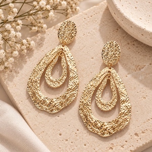 May include: A pair of gold-toned drop earrings featuring a textured, hammered metal finish. Each earring consists of a circular stud connected to a large, open teardrop shape that frames a smaller, central teardrop element, creating a layered look.