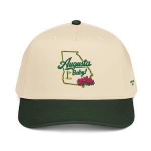 Può includere: Un cappellino da baseball color crema con visiera verde scuro. Il cappellino presenta un disegno ricamato dello stato della Georgia in verde e oro, con la scritta "Augusta Baby!" e una bandiera da golf. Sono ricamati anche fiori rosa.