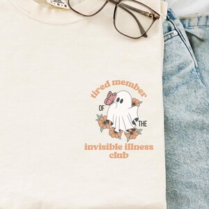 Könnte beinhalten: Cremefarbenes T-Shirt mit dem Text "tired member of the invisible illness club" und einer Geistergrafik mit einem Schmetterling. Das Shirt hat einen Rundhalsausschnitt und kurze Ärmel. Eine Brille liegt auf dem Shirt.