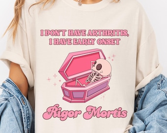 May include: Cream-colored t-shirt with the text "I DON'T HAVE ARTHRITIS, I HAVE EARLY ONSET" above a pink coffin with a skeleton inside. The word "Rigor Mortis" is written below the coffin in pink script.