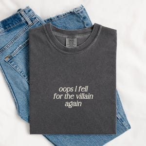 May include: A dark gray t-shirt with the text "oops I fell for the villain again" in white, paired with blue denim jeans. The t-shirt is folded neatly, and the jeans are partially visible.
