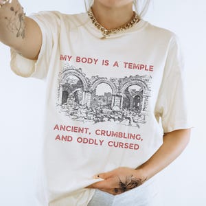 May include: Cream-colored t-shirt with the text "MY BODY IS A TEMPLE" above a black and white illustration of ancient ruins. Below the illustration, the text reads "ANCIENT, CRUMBLING, AND ODDLY CURSED".