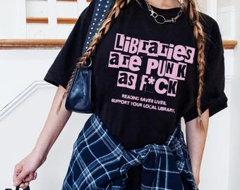 May include: Black t-shirt with a pink graphic that reads "Libraries are Punk as F*CK". The text is in a distressed font style. The shirt also has the text "Reading Saves Lives. Support Your Local Library." printed below the graphic.