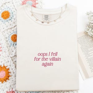 May include: Cream-colored t-shirt with the phrase "oops I fell for the villain again" in maroon lettering. A delicate butterfly necklace rests on the shirt. The shirt is on a white surface with a crocheted blanket and a book.