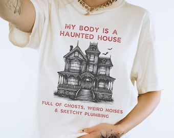 May include: Cream-colored t-shirt with the text "MY BODY IS A HAUNTED HOUSE" in red above a detailed black and white illustration of a spooky mansion. Below the house, the text reads "FULL OF GHOSTS, WEIRD NOISES & SKETCHY PLUMBING."