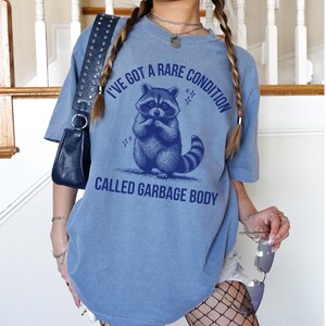 May include: A blue t-shirt with the text "I'VE GOT A RARE CONDITION CALLED GARBAGE BODY" and a raccoon graphic. The raccoon is in a seated position with its hands clasped. The shirt has short sleeves.