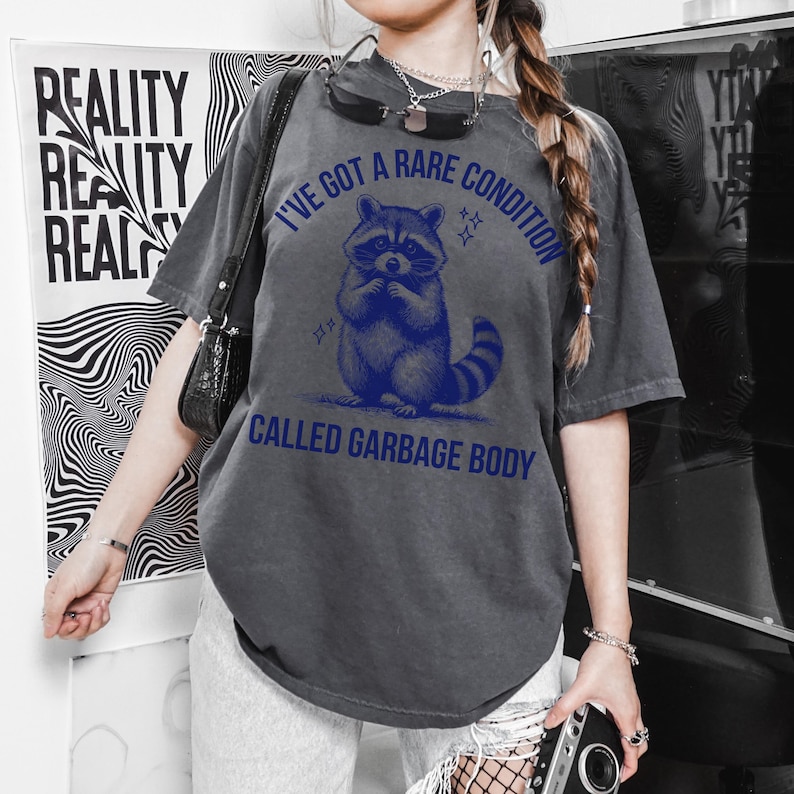 May include: Dark gray t-shirt with a blue graphic of a raccoon and the text "I'VE GOT A RARE CONDITION CALLED GARBAGE BODY". The shirt is a relaxed fit, with short sleeves.