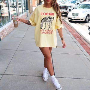 May include: A light yellow t-shirt with the text "IT'S NOT RARE, IT'S RARELY DIAGNOSED" and a zebra graphic. The shirt is oversized and worn with white sneakers.