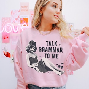 Può includere: Felpa rosa chiaro con la scritta "TALK GRAMMAR TO ME" e un'illustrazione in bianco e nero di una donna. La felpa ha maniche lunghe e girocollo.