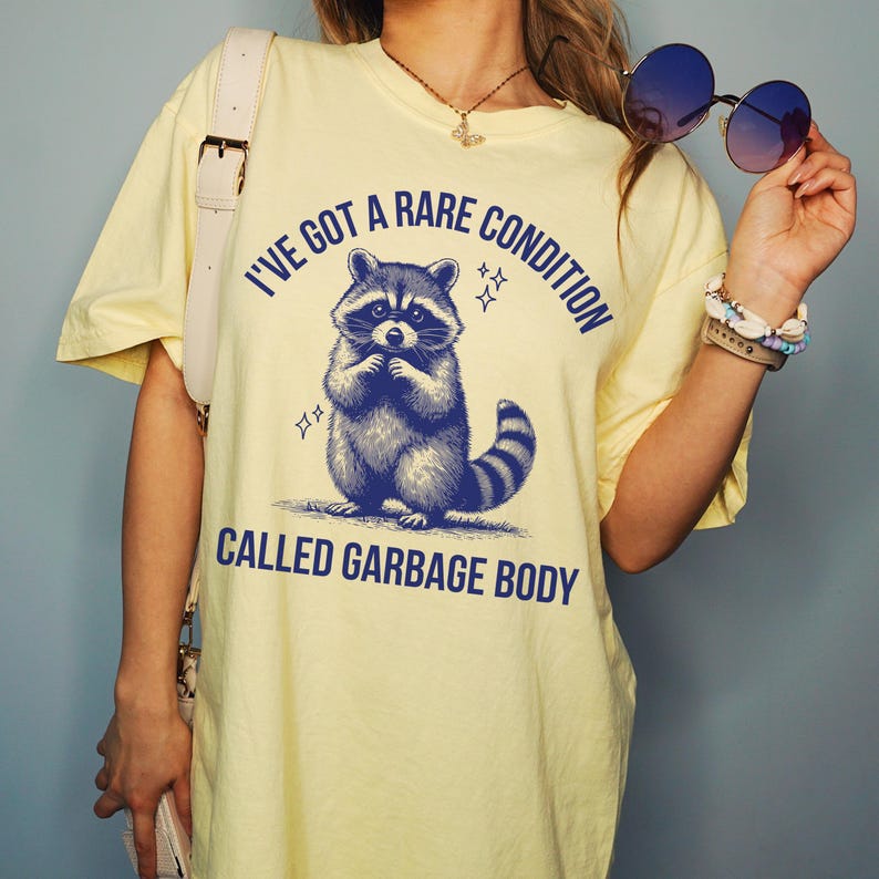 May include: A light yellow t-shirt with a blue graphic of a raccoon and the text "I'VE GOT A RARE CONDITION CALLED GARBAGE BODY". The raccoon is standing with its paws together. The shirt has short sleeves.