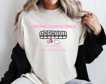 May include: Cream-colored t-shirt with the text "CHRONIC ILLNESS GIRLIE" in pink. Below the text is a graphic of a pill organizer with pills and the words "low energy, high maintenance."