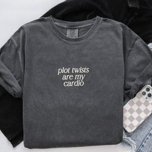Pode incluir: Camisola cinza escuro com gola redonda com o texto branco "plot twists are my cardio". A camisola tem um aspeto vintage e lavado. Um telemóvel com uma capa xadrez está ao lado.