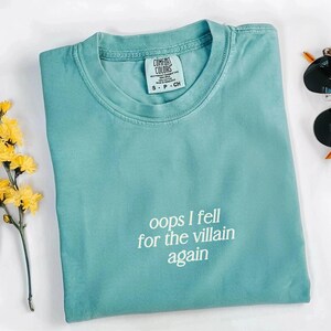May include: A turquoise Comfort Colors t-shirt with the text "oops I fell for the villain again" in white. The shirt has a crew neck and short sleeves. A small tag is visible near the collar. Yellow flowers and sunglasses are in the background.