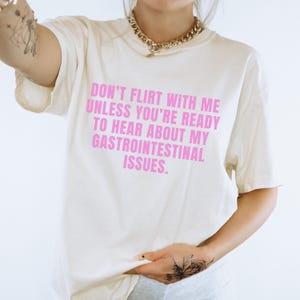 May include: Cream-colored t-shirt with the text "DON'T FLIRT WITH ME UNLESS YOU'RE READY TO HEAR ABOUT MY GASTROINTESTINAL ISSUES" in pink. The shirt has short sleeves and a relaxed fit.
