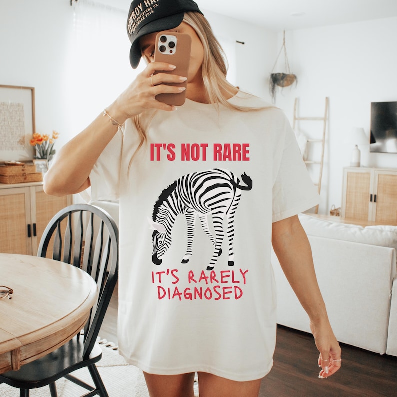 May include: Cream-colored t-shirt with the text "IT'S NOT RARE" in red above a black and white zebra illustration. Below the zebra, the text "IT'S RARELY DIAGNOSED" is also in red. The shirt is a casual, oversized fit.