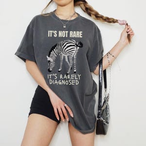 May include: Dark grey t-shirt with a zebra graphic and the text "IT'S NOT RARE, IT'S RARELY DIAGNOSED". The zebra is black and white. The shirt is a casual, oversized fit.