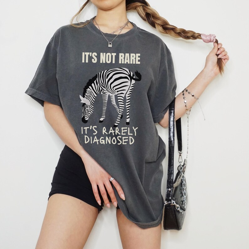 May include: Dark grey t-shirt with a zebra graphic and the text "IT'S NOT RARE, IT'S RARELY DIAGNOSED". The zebra is black and white. The shirt is a casual, oversized fit.